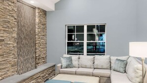 A modern living room with a stone accent wall, a sectional sofa with patterned cushions, and a large window with a view of the city. - Howard Johnson by Wyndham Middletown Newport Area (Middletown)