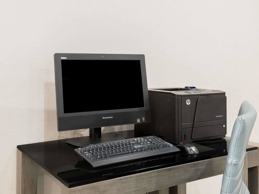 Un écran Lenovo, une imprimante HP et un clavier sont posés sur un bureau en bois, devant une chaise bleu clair.