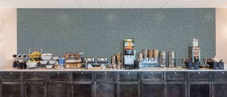 A breakfast bar with various food items and beverages, a coffee machine, and storage cabinets.