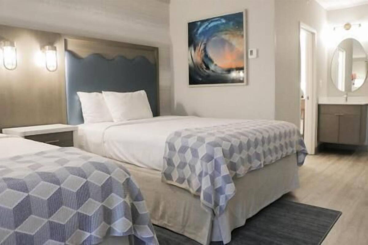 A hotel room with two beds, a painting on the wall, a bathroom with a round mirror, and a small desk.