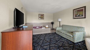 Ein Hotelzimmer mit einem Flachbildfernseher auf einem Holztisch, einem Sofa, einem kleinen Tisch mit einer Lampe, einer Badewanne und einem gemusterten Teppich.