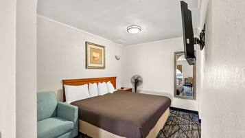 A hotel room with a bed, a chair, a bedside table, a fan, and a mirror.