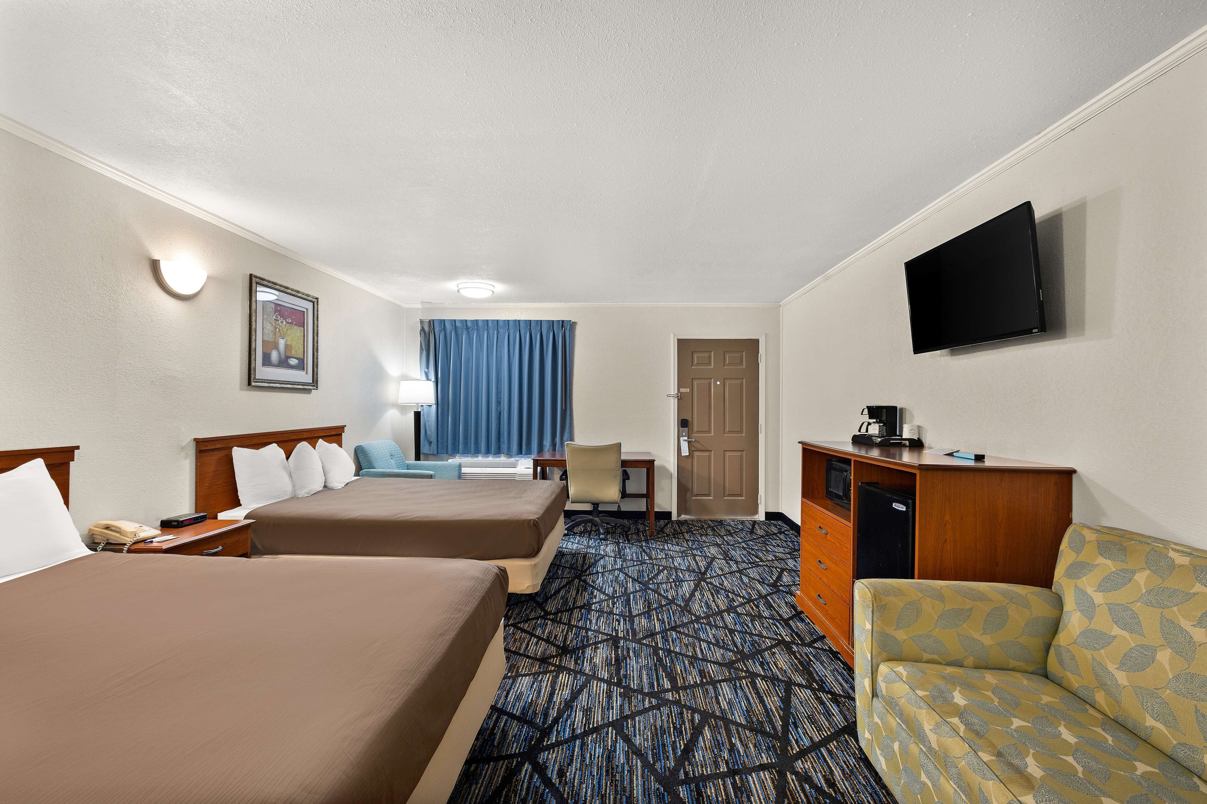 Habitación de hotel con dos camas, un sofá, un escritorio con computadora, televisor y alfombra estampada.