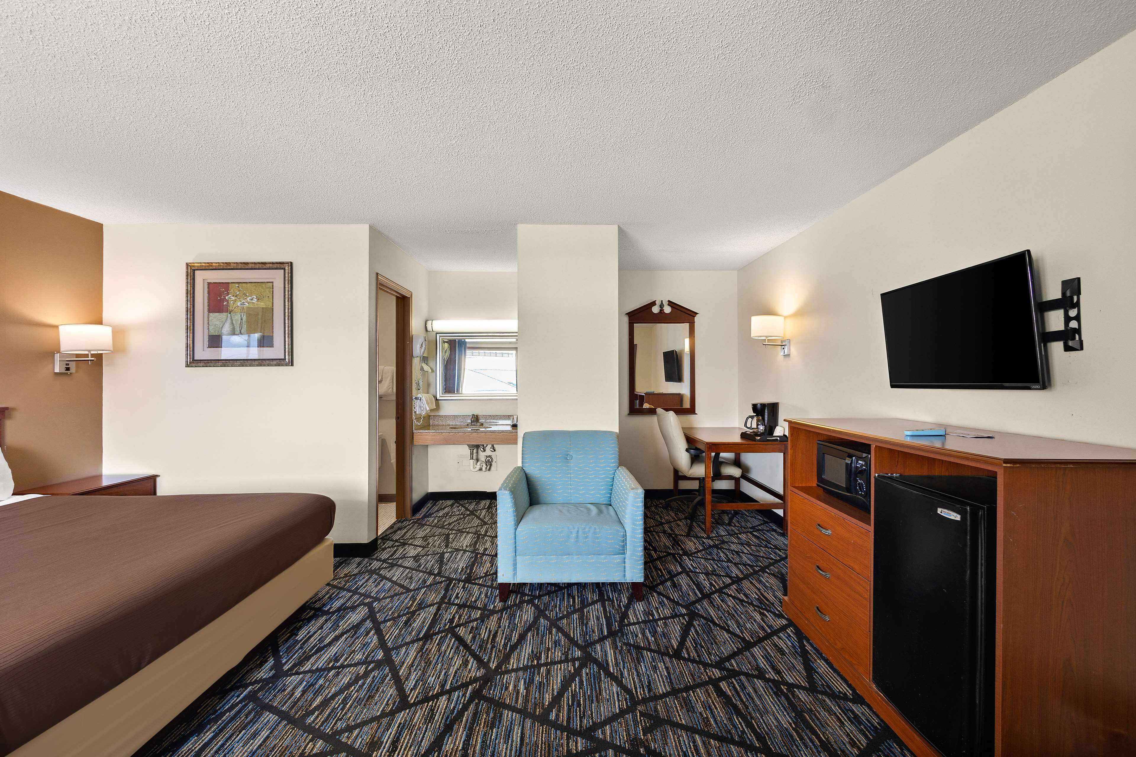 A hotel room with a bed, a desk with a chair, a microwave, a refrigerator, and a television on the wall.