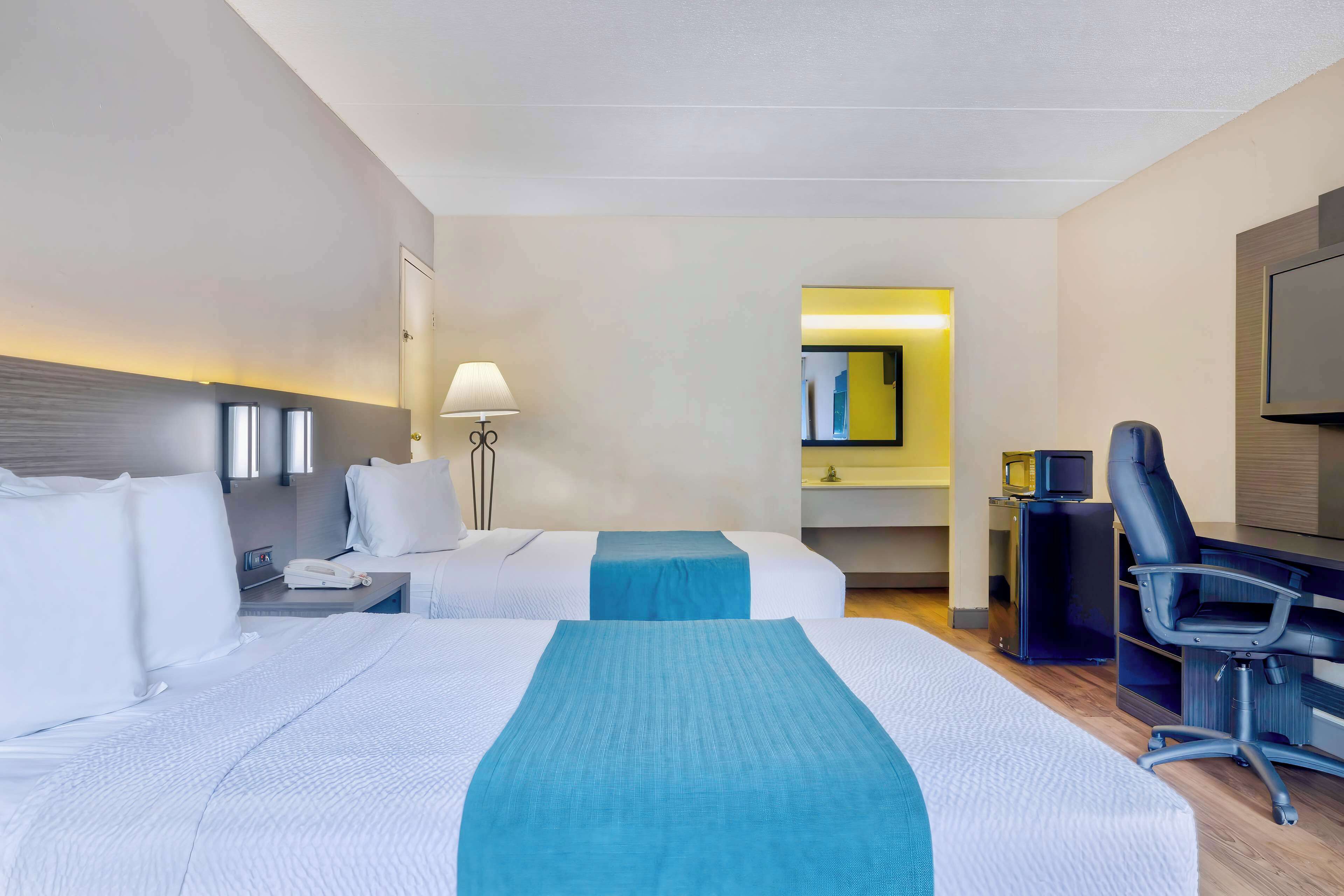 A hotel room with two beds, a desk, a chair, a TV, and a lamp.
