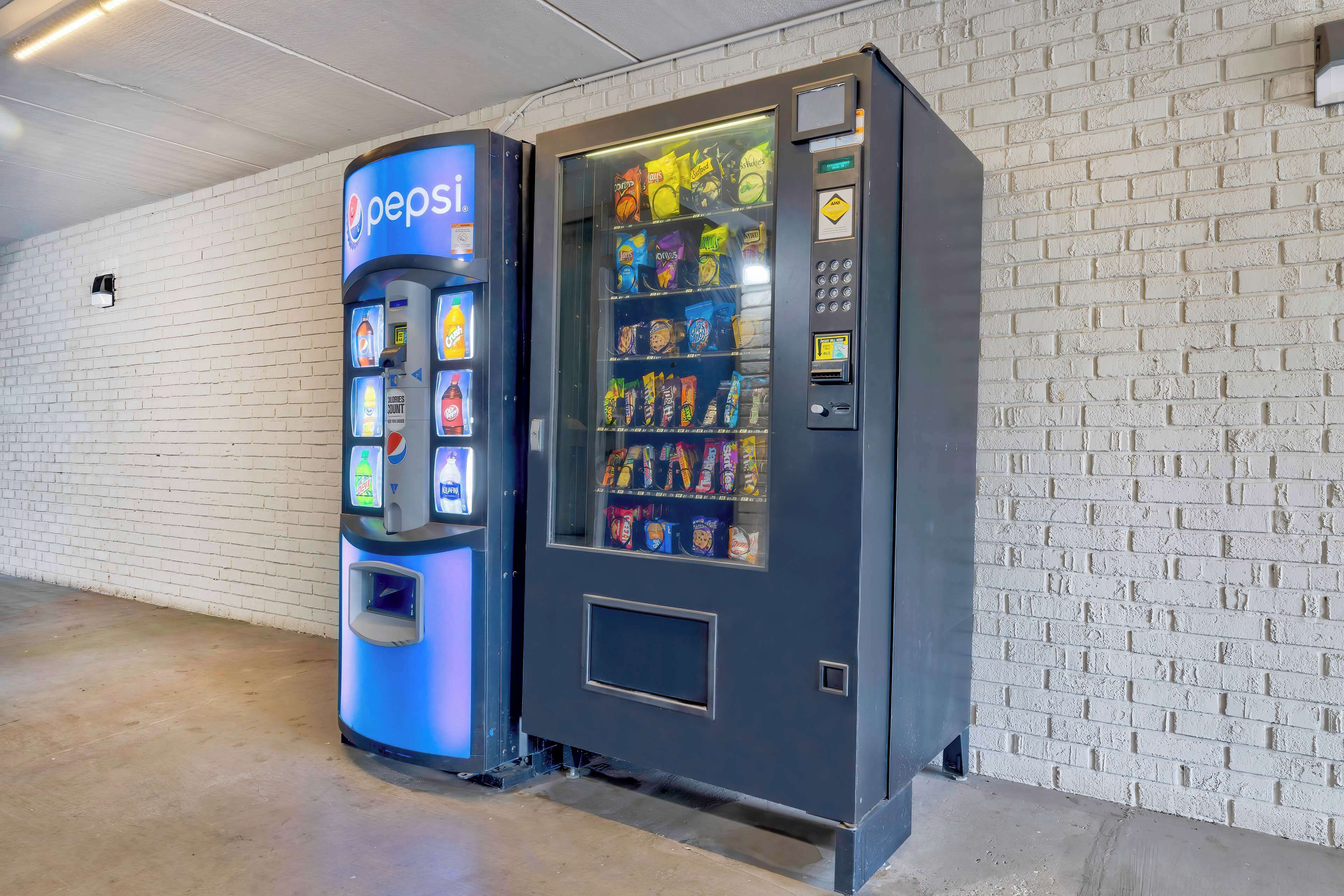 A vending machine with a Pepsi logo and a snack vending machine next to a white brick wall.
