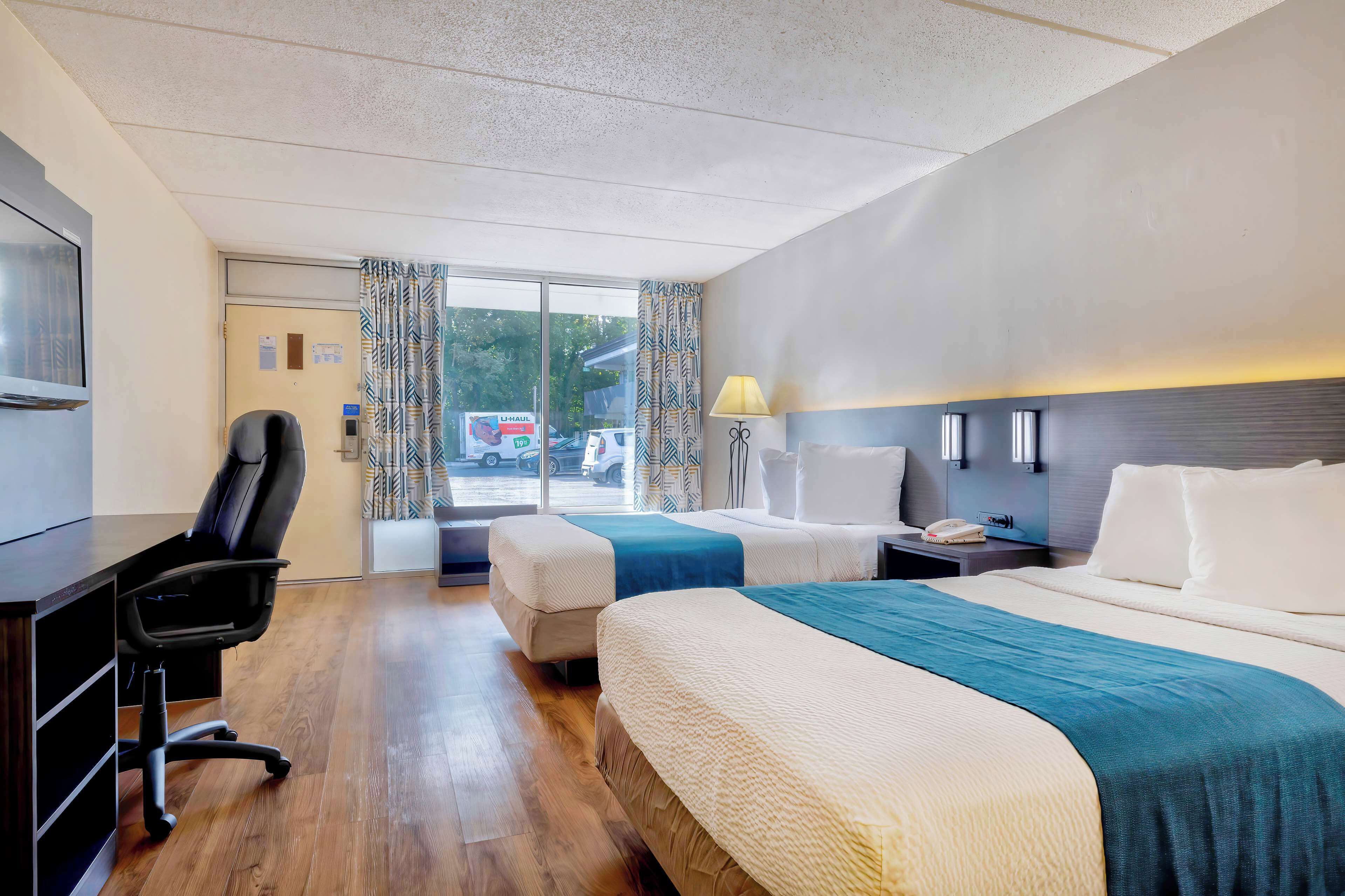 A hotel room with two beds, a desk, a chair, a lamp, and a view of the outside.