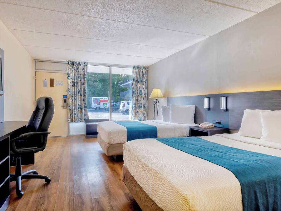 A hotel room with two beds, a desk, a chair, a lamp, and a view of the outside.