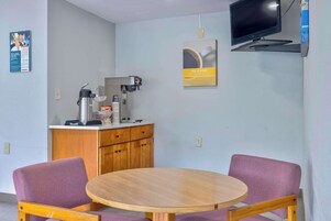 A small room with a round wooden table, two purple chairs, a coffee machine, a TV mounted on the wall, and a poster advertising coffee.