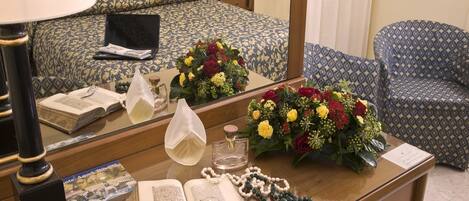 A hotel room with a bed, a desk with a book, a lamp, a mirror, and a bouquet of flowers.