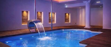 A swimming pool with a waterfall feature, wooden flooring, and wall-mounted light fixtures.