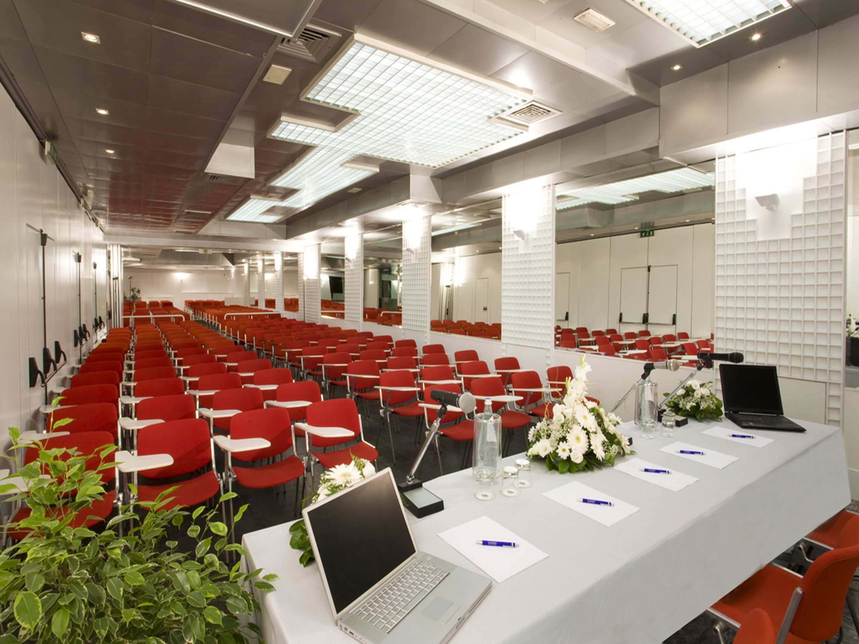 Una sala conferenze con file di sedie rosse, un lungo tavolo con portatili, microfoni e composizioni floreali.