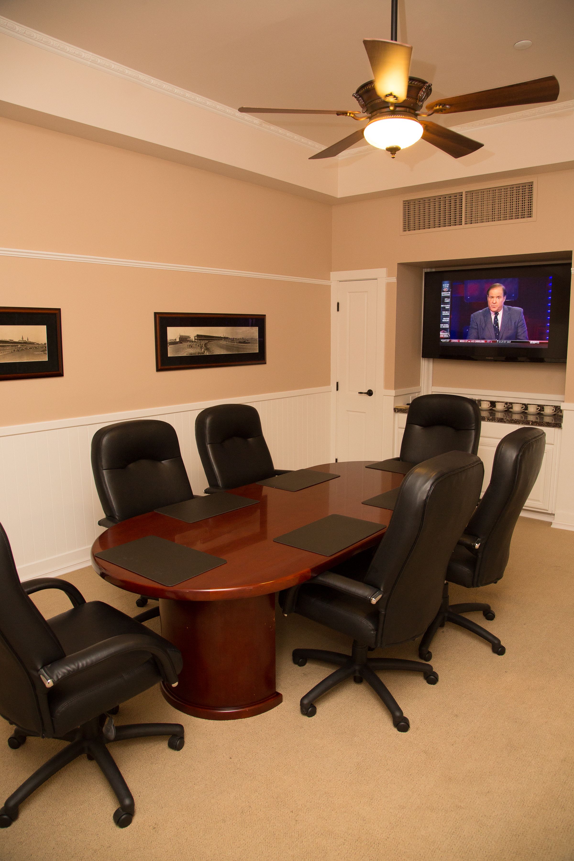 A conference room with a wooden table, black chairs, a ceiling fan, framed pictures on the wall, and a television screen.