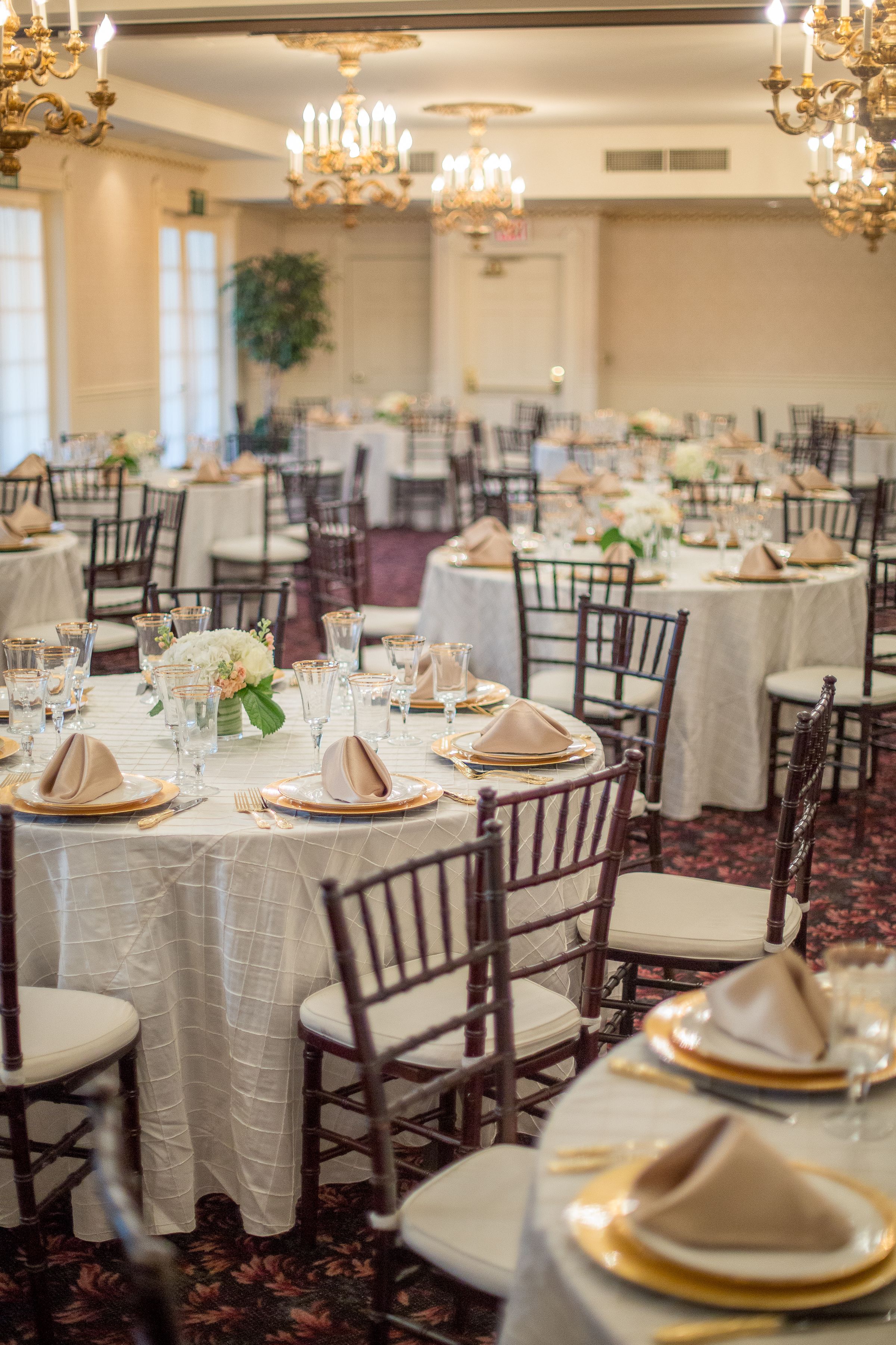 A banquet hall with round tables set for a formal event, featuring white tablecloths, gold-rimmed plates, and elegant floral centerpieces.