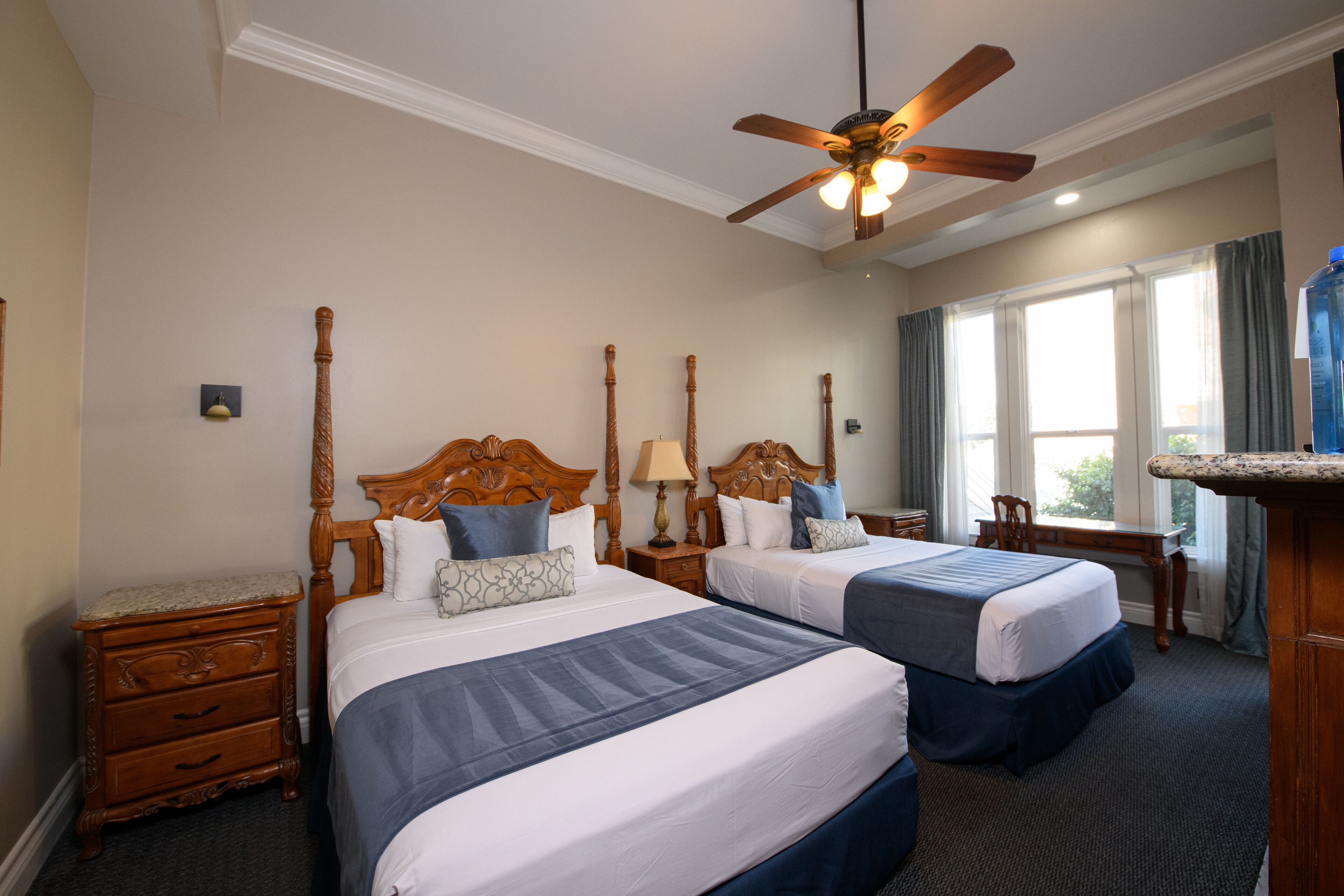 A hotel room with two beds, a wooden headboard, a desk, and a large window with curtains.
