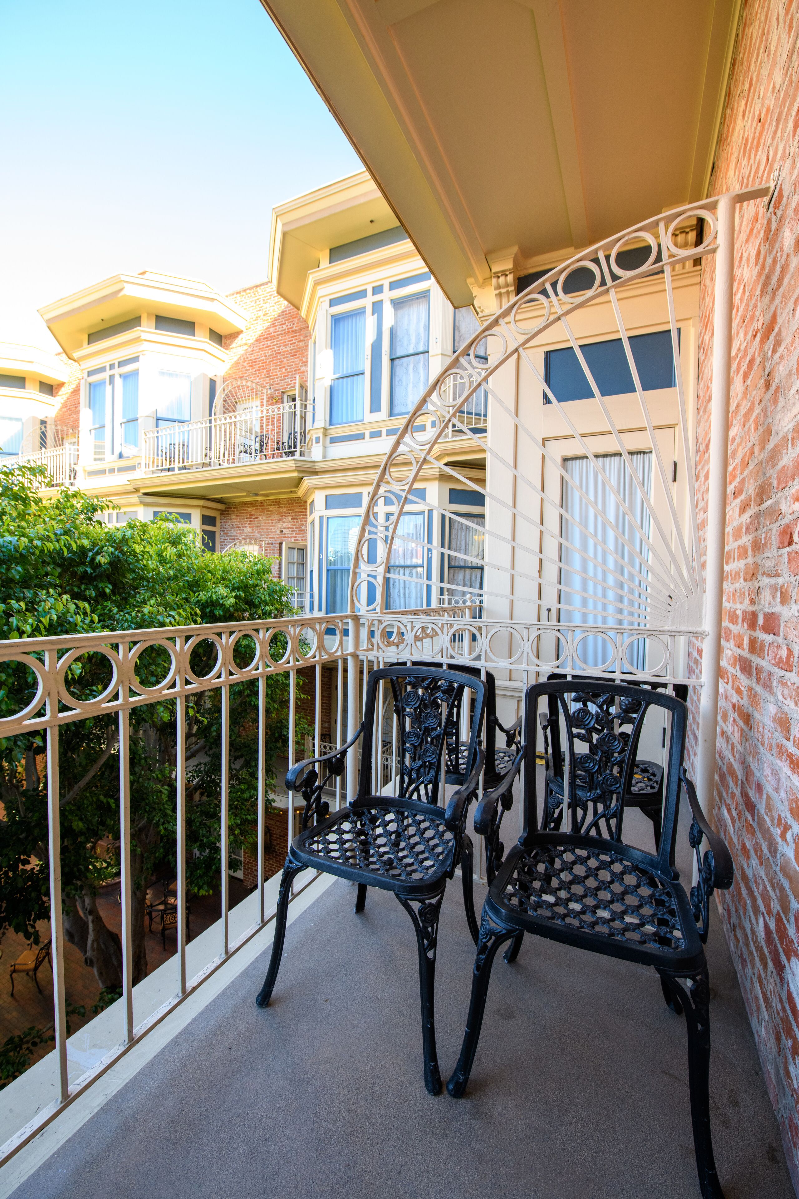 A balcony with two ornate metal chairs, a white railing, and a view of a brick building with multiple windows and balconies.