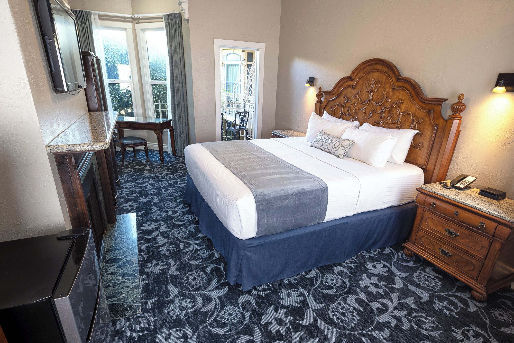 A hotel room with a large bed, a desk, and a chair. There is a window with curtains, a door, and a view of a building outside.