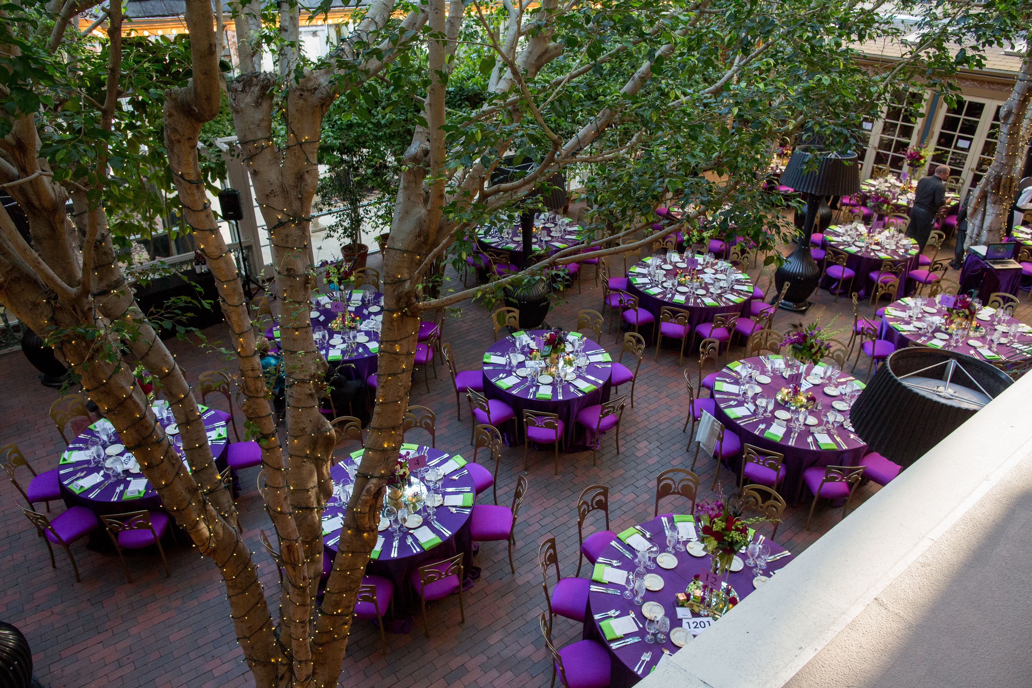 Un espace de restauration en plein air avec des tables rondes et des chaises violettes, entouré d’arbres et doté d’un sol en briques.