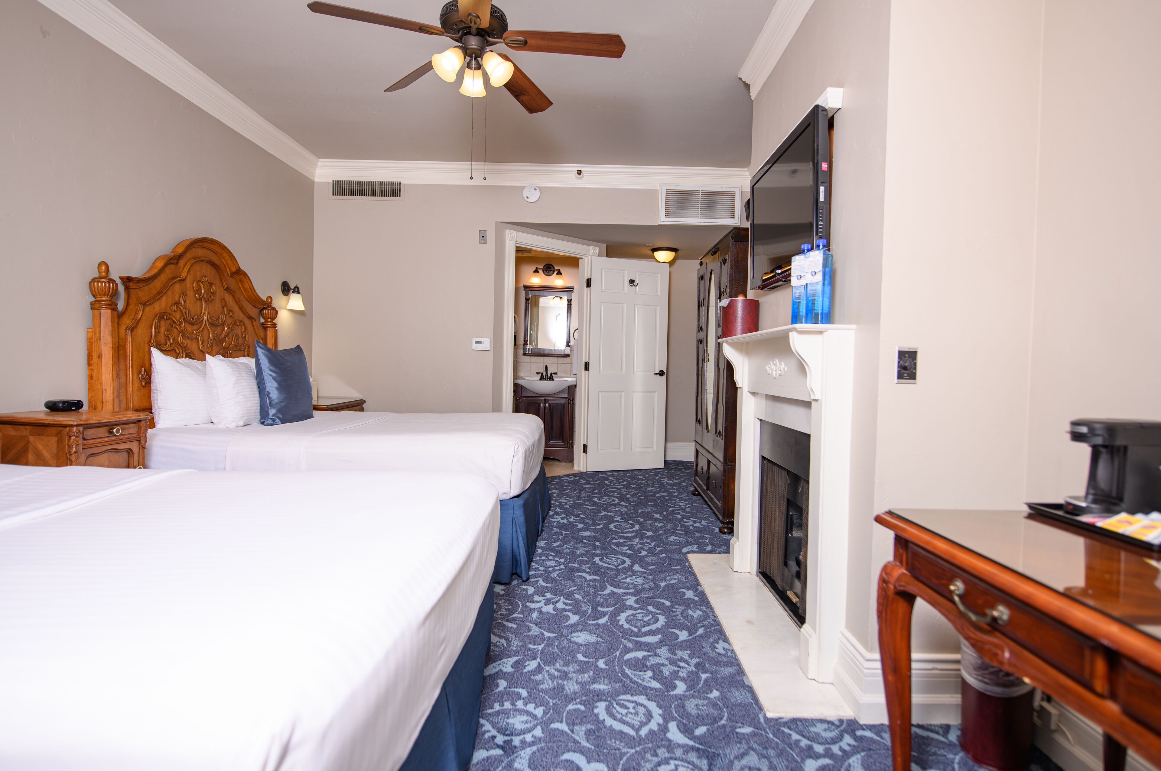 A hotel room with two beds, a ceiling fan, a fireplace, and a TV.