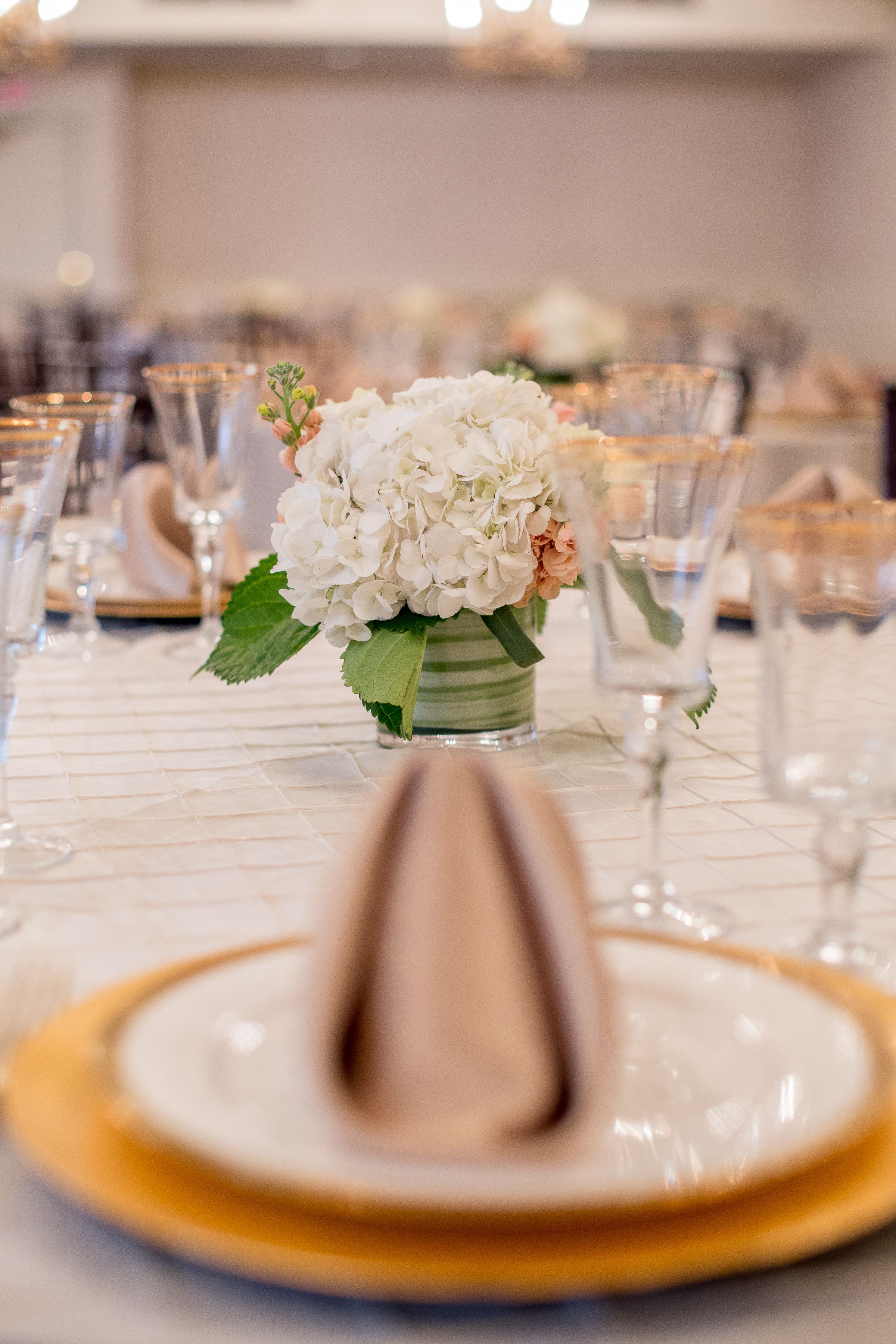 Una mesa vestida con mantel blanco, un jarrón con flores blancas y un plato hondo dorado.