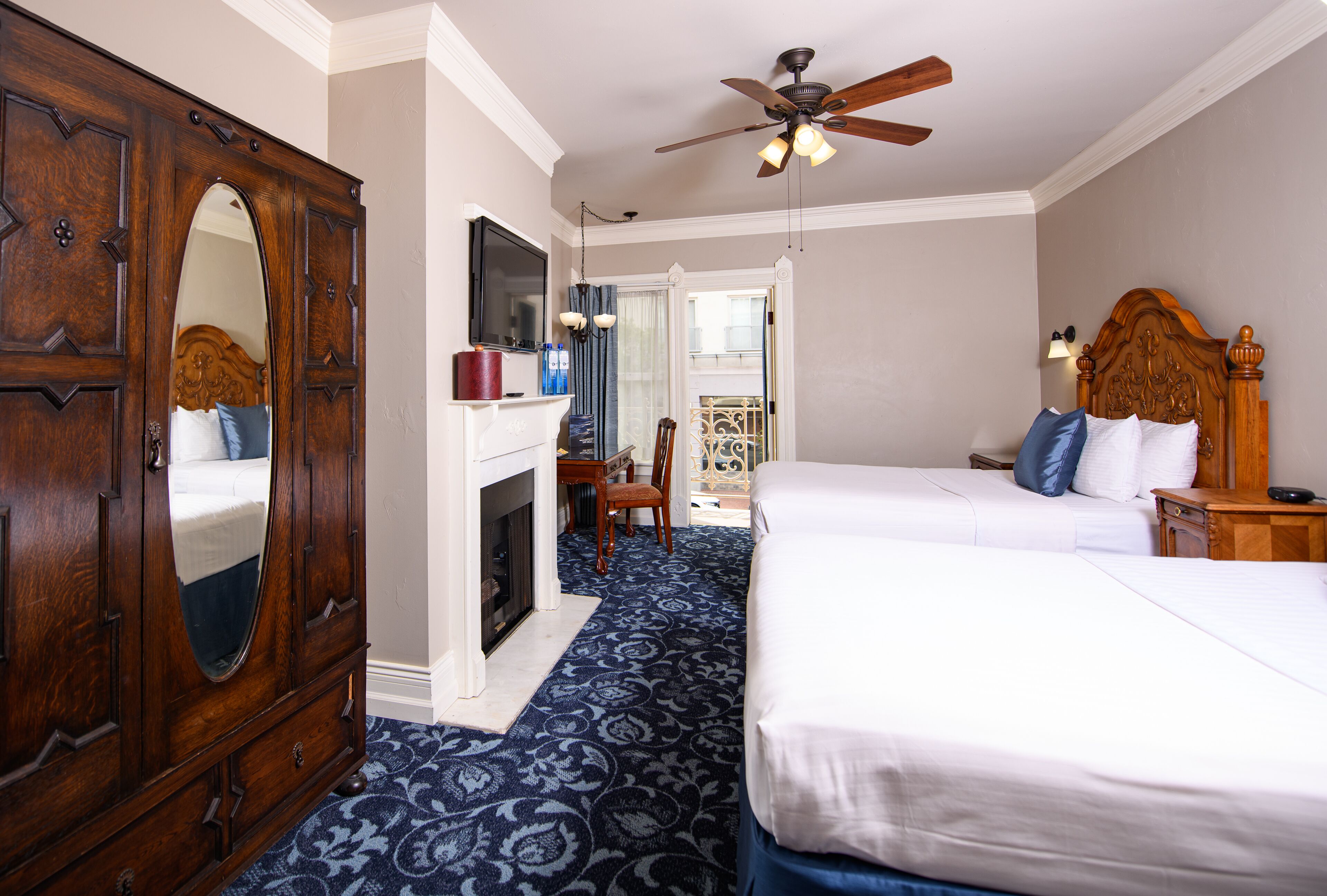A hotel room with a large wooden wardrobe, a fireplace, two beds, a ceiling fan, and a balcony with a view.