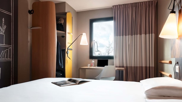 A hotel room with a bed, a desk, a chair, a window with curtains, and a lamp.