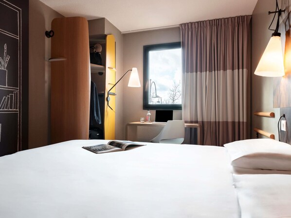 A hotel room with a bed, a desk, a chair, a window with curtains, and a lamp.