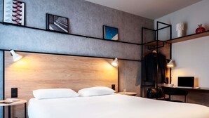 A modern hotel room with a large bed, bedside tables, a desk with a laptop, and wall-mounted shelves.