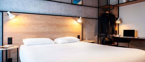 A modern hotel room with a large bed, bedside tables, a desk with a laptop, and wall-mounted shelves.