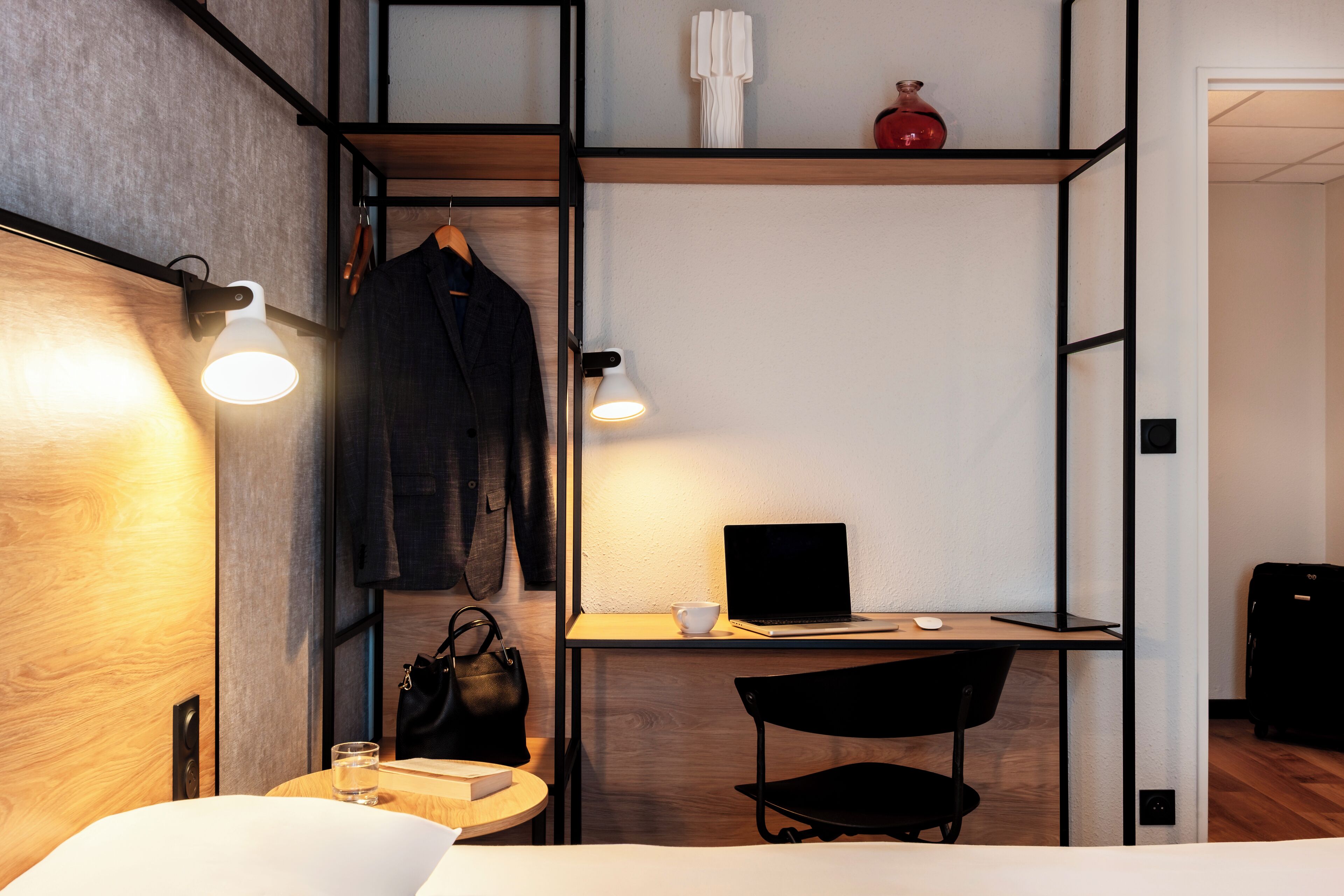 A compact hotel room with a bed, a desk with a laptop, a chair, a small table with a glass, and a hanging coat.