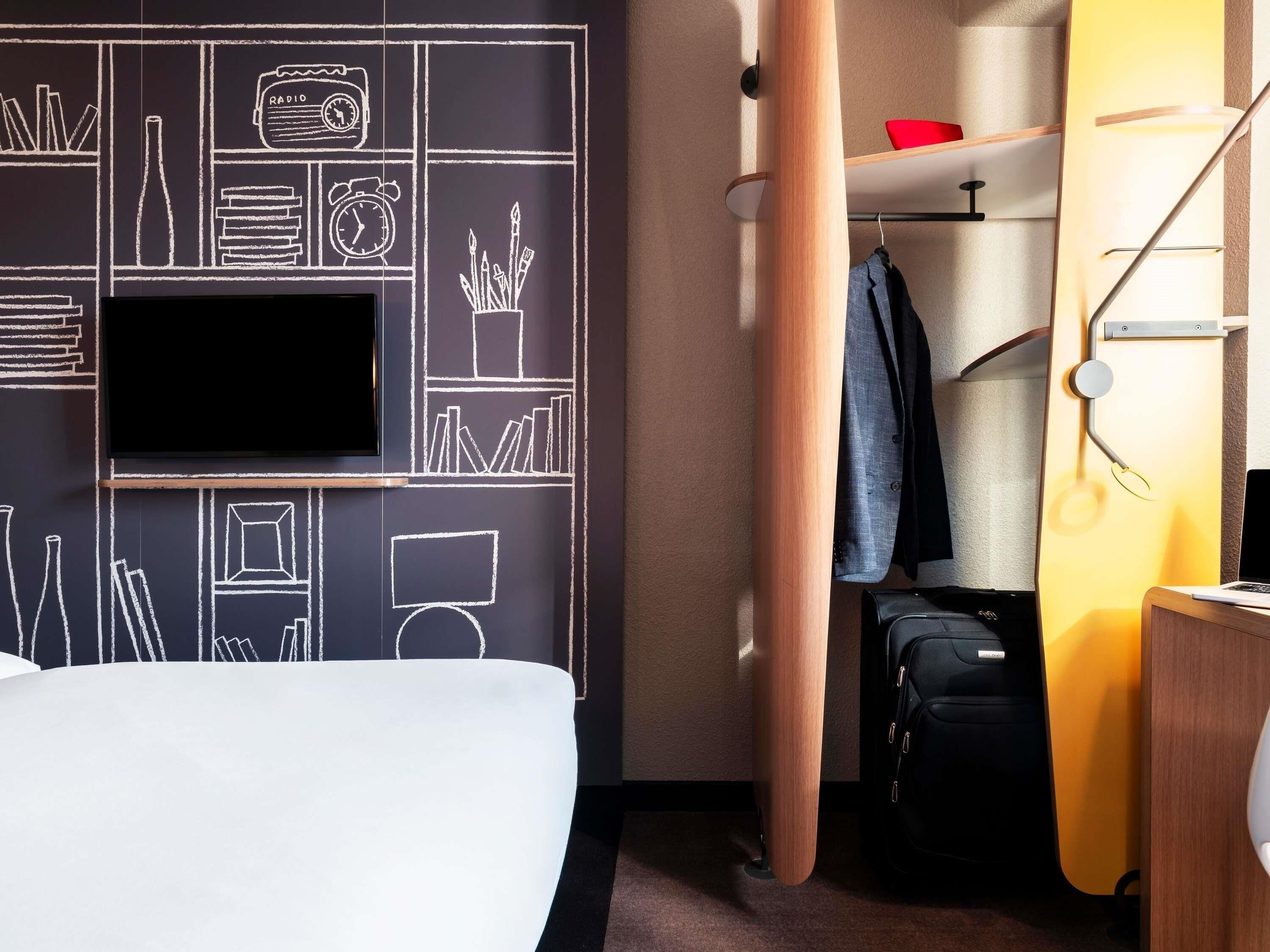 A hotel room with a bed, a television, a wardrobe, and a desk with a lamp.