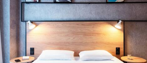 A modern hotel room with a large bed, two bedside tables, a wooden headboard, and framed artwork on the wall.
