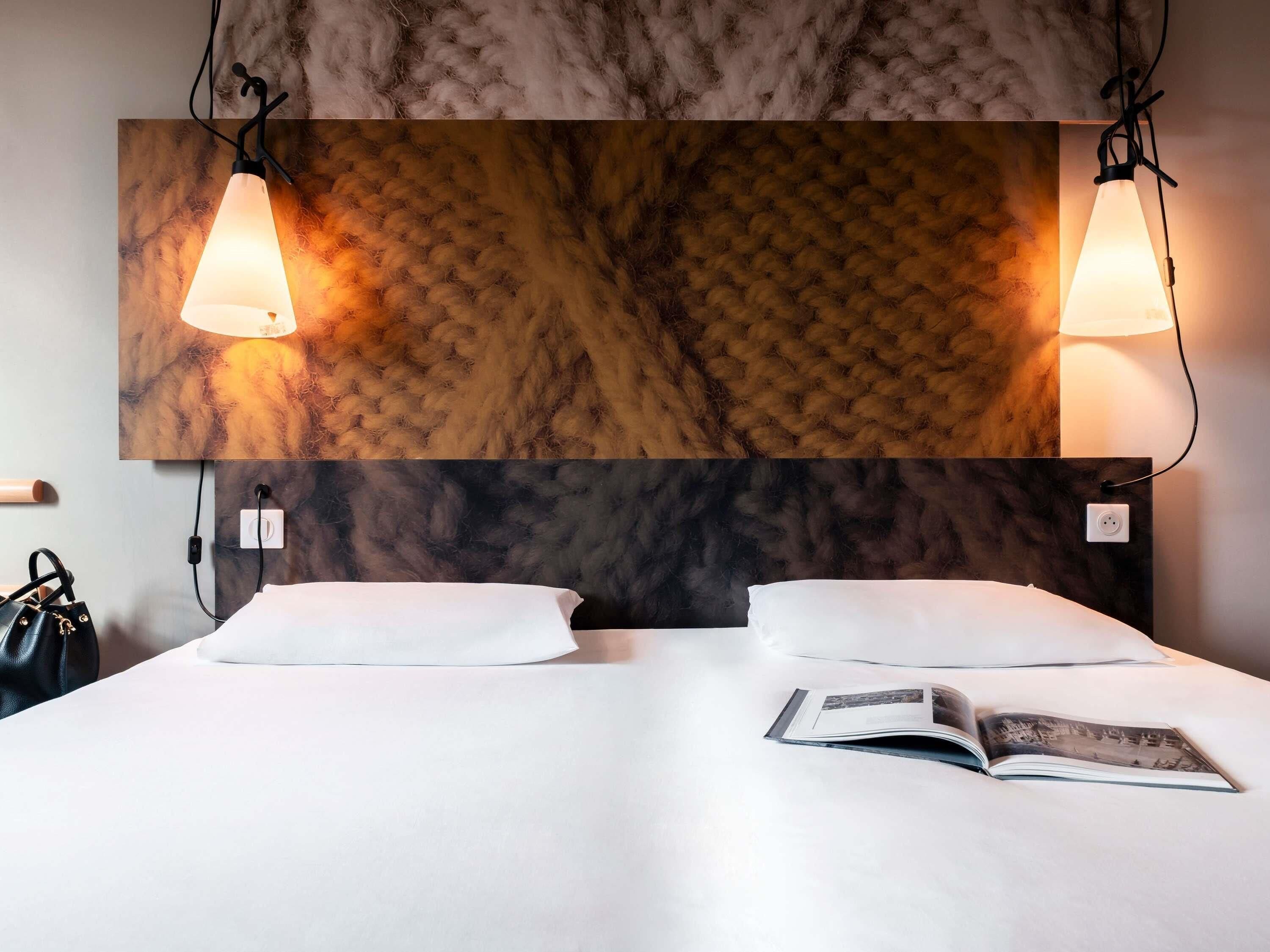 Habitación de hotel con cama, dos lámparas de noche, un aplique de pared y una revista sobre la cama.