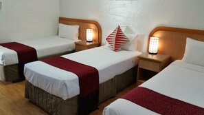 Habitación de hotel con tres camas individuales, cada una con cabecero de madera, ropa de cama blanca y una manta roja. Hay mesitas de noche con lámparas y un cojín rojo y blanco en una cama.