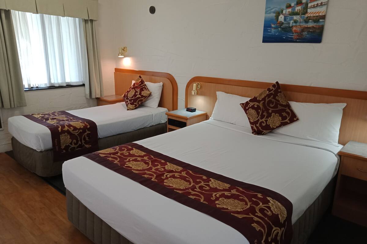 Ein Hotelzimmer mit zwei Betten, jeweils mit einem hölzernen Kopfteil, weißer Bettwäsche und einer Bettdecke mit rotem und goldenem Muster. Ein Nachttisch mit Lampe, ein Fenster mit Vorhängen und ein Gemälde einer Küstenlandschaft sind ebenfalls vorhanden.