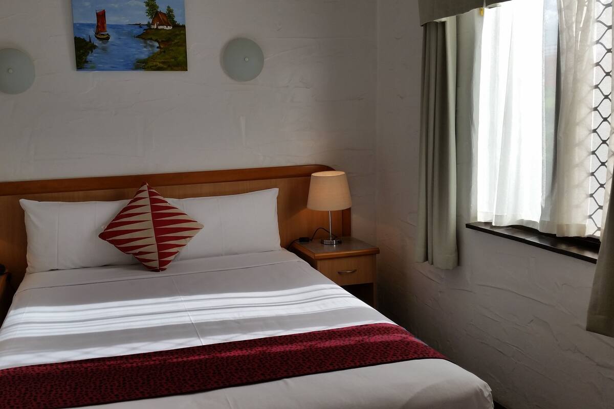 Ein Hotelzimmer mit Bett, zwei Nachttischen mit Lampen, einem Holzkopfstück und einem roten und weißen Kissen. An der Wand hängt ein Bild.