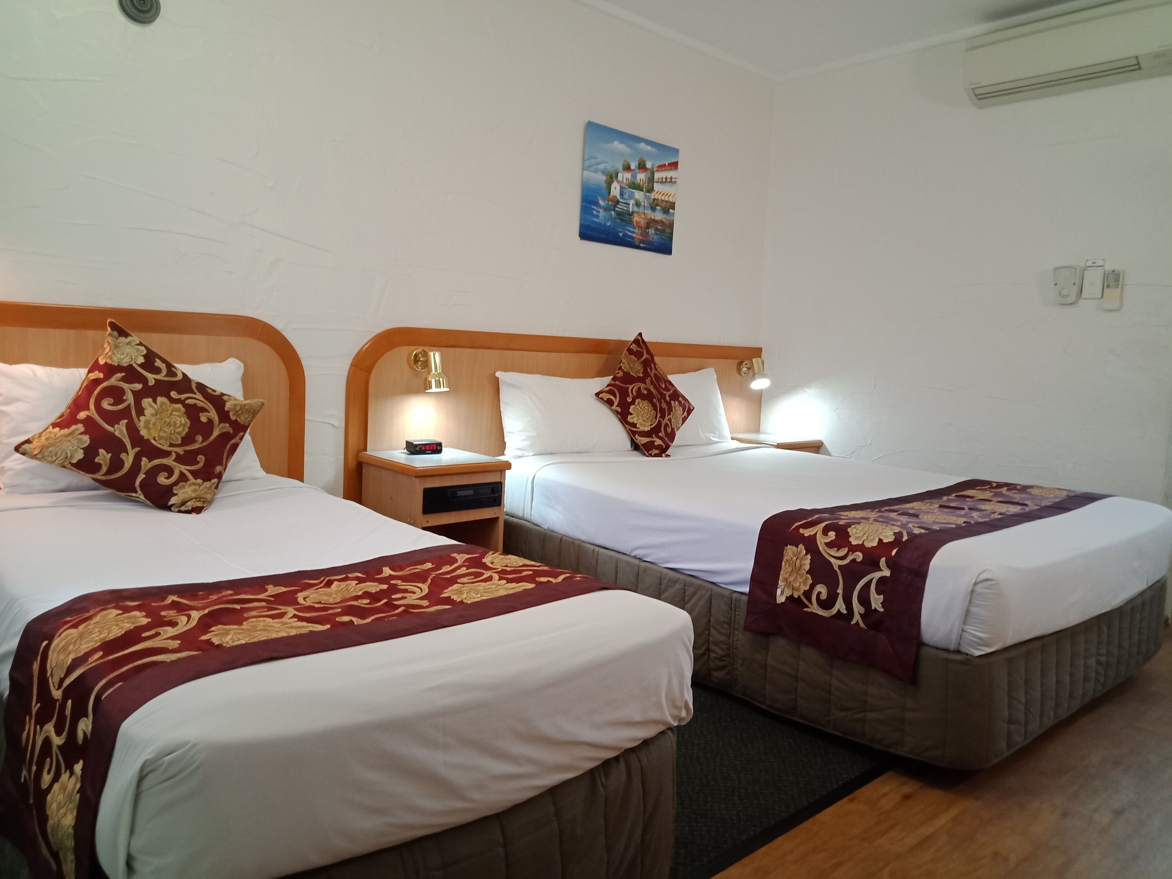 A hotel room with two beds, each with a wooden headboard and bedside tables. There is a painting on the wall above the beds.