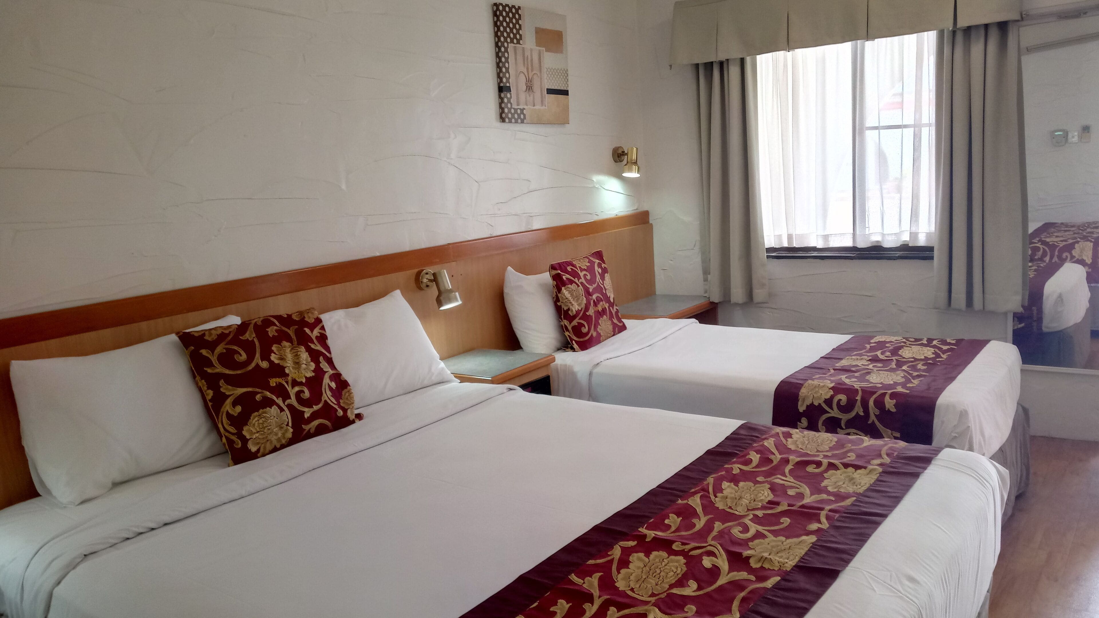 A hotel room with two beds, each with white linens and maroon decorative pillows. There is a wall-mounted artwork above the beds and a window with curtains to the right.