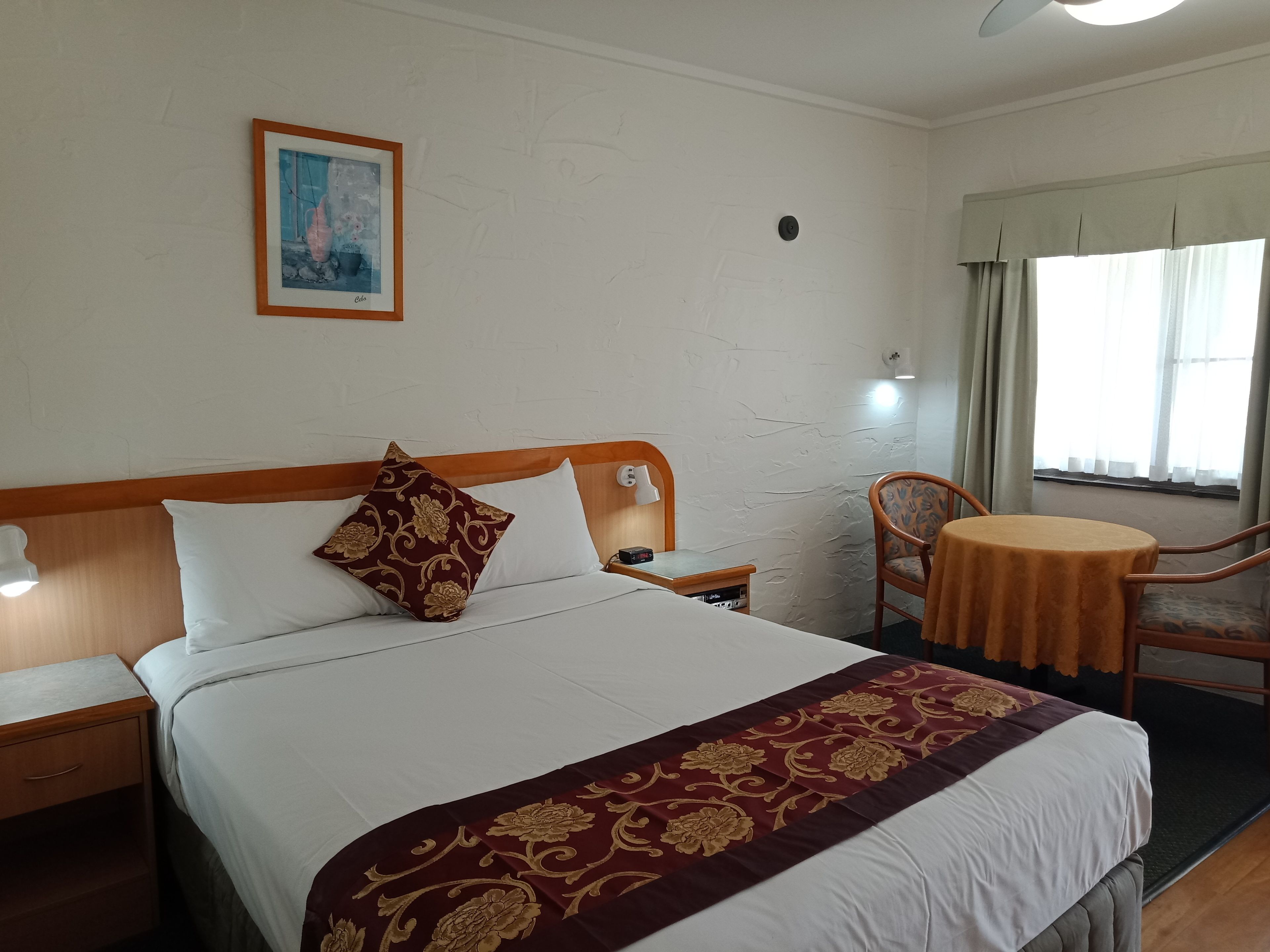A hotel room with a bed, a nightstand, a chair, a small table, a painting on the wall, and a window with curtains.