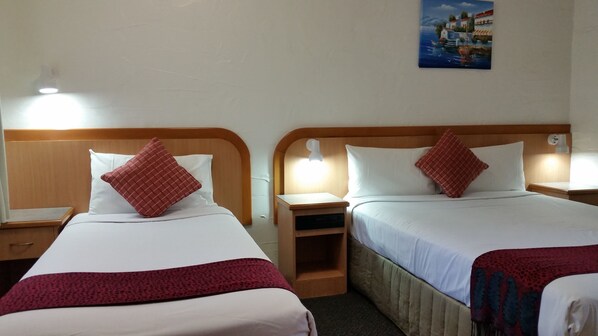 Habitación de hotel con dos camas, cada una con cabecero de madera, ropa de cama blanca y almohadas rojas a cuadros. Hay una mesita de noche con lámpara a cada lado de las camas, y un pequeño estante entre ellas.