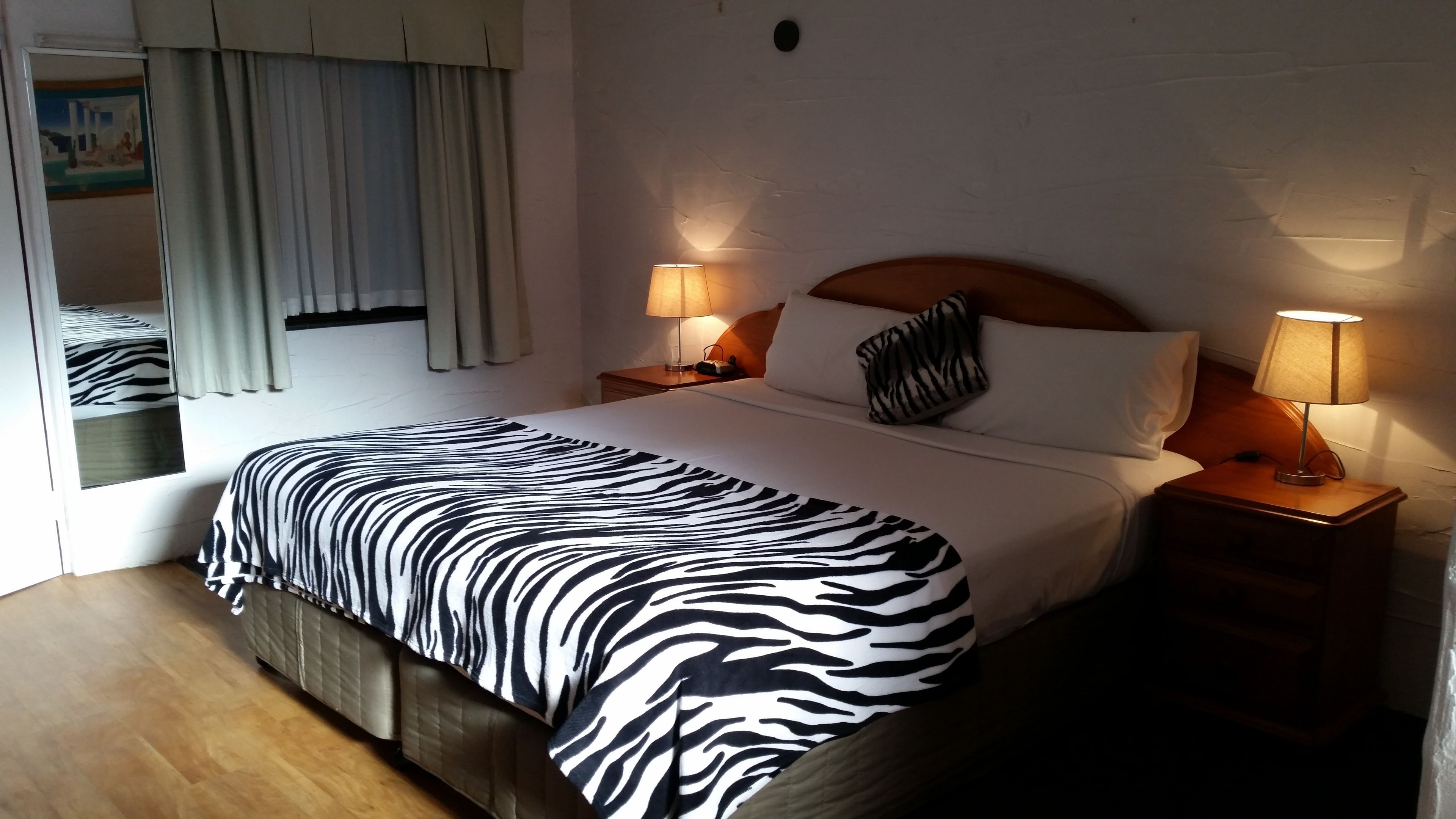 Habitación de hotel con edredón estampado de cebra, dos lámparas de noche, cabecera de madera y una ventana con cortinas.
