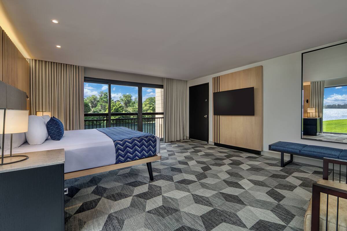 Une chambre d’hôtel comprenant un lit, un bureau avec une lampe, une télévision, un banc et une grande fenêtre donnant sur un paysage verdoyant et un plan d’eau.