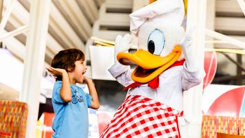 Une personne vêtue d’un costume à carreaux, portant un grand bec et un chapeau de chef, interagissant avec un enfant qui porte un t-shirt bleu.