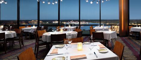 Ein Restaurant mit gedeckten Tischen, großen Fenstern mit Blick auf die Stadtlandschaft und modernen Kronleuchtern.