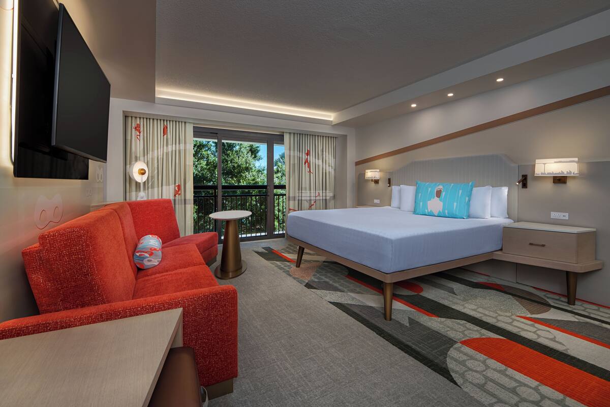 Une chambre d’hôtel avec un lit, un fauteuil rouge, une petite table, une table de chevet et un téléviseur fixé au mur.
