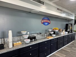 Zona de desayuno tipo bufé con un letrero que dice 'Baymont Breakfast Corner', diversas opciones de comida y una máquina de café.