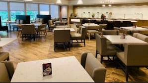 Un comedor moderno con varias mesas y sillas, una cocina al fondo y grandes ventanales que permiten la entrada de luz natural.