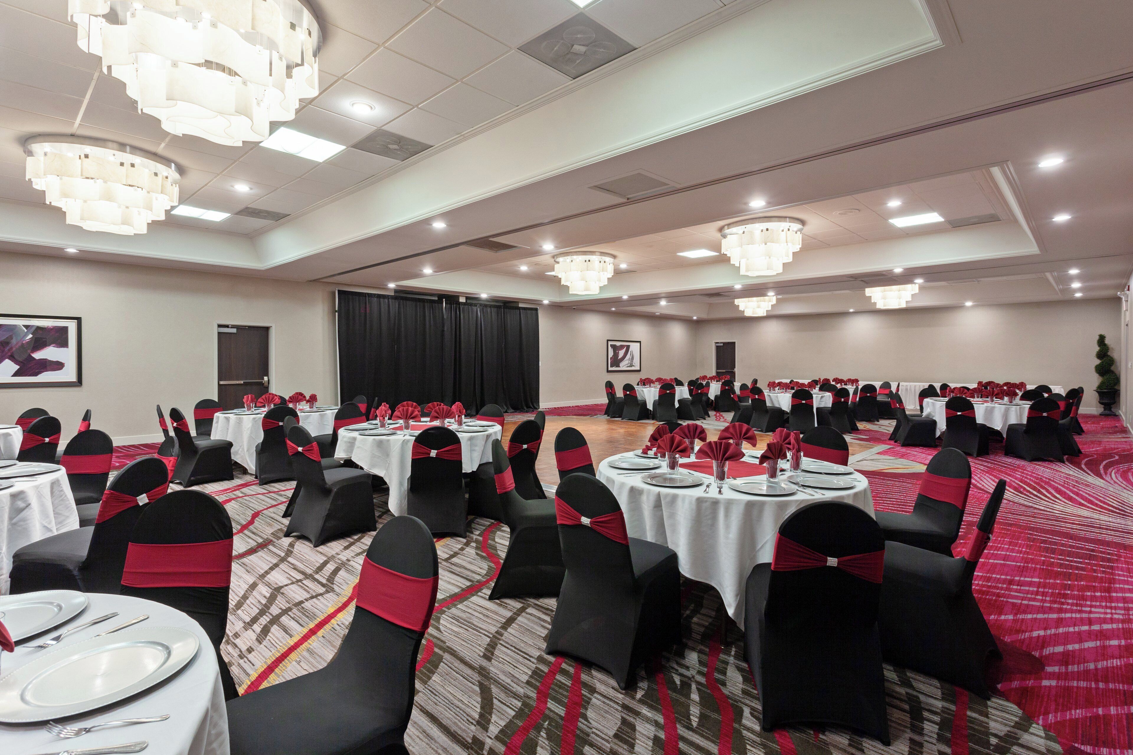 A banquet hall with round tables, black chairs with red sashes, and chandeliers.