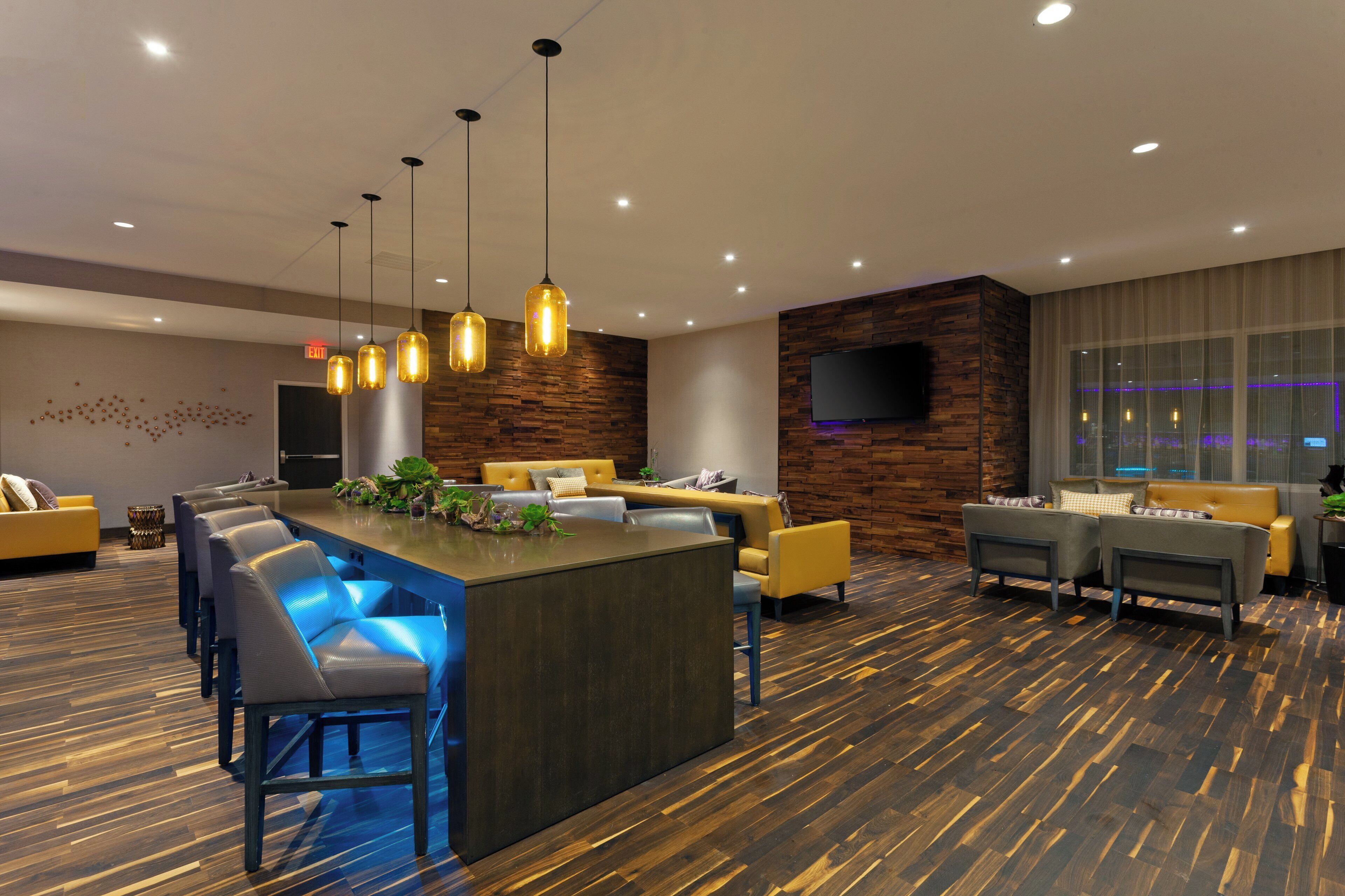 A modern hotel lobby with a central bar area, seating arrangements, and a large television screen.