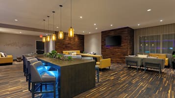 A modern hotel lobby with a central bar area, seating arrangements, and a large television screen.